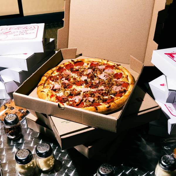 A Saruzzo's New York Pizzeria meat lover's pizza in an open box, surrounded by other pizza boxes and spice shakers.