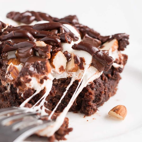 A rich chocolate brownie with melted marshmallows, chocolate drizzle, and almonds, with a fork taking a bite.
